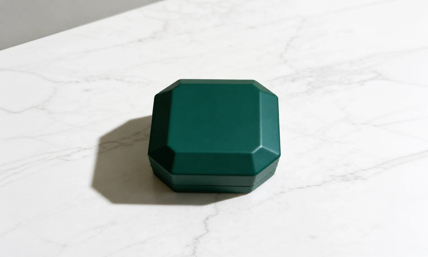 Closed deep-green faceted jewelry box on white marble under directional natural light.