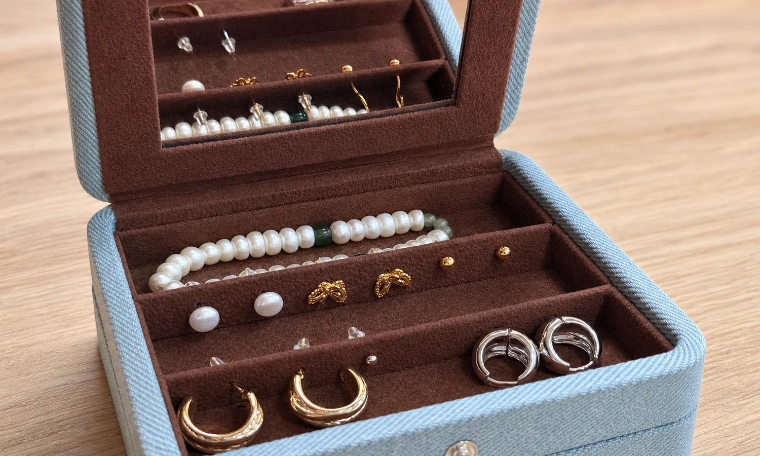 Open blue fabric jewelry organizer with brown divided inserts holding pearl strands, earrings, and hoops.