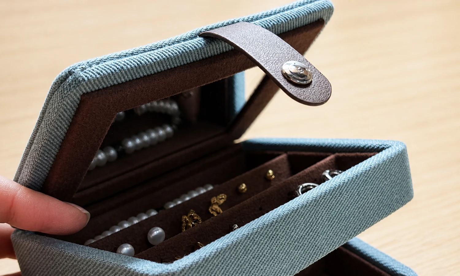 Close view of a blue woven jewelry organizer with brown lining, a leather-like strap, and a metal snap closure.
